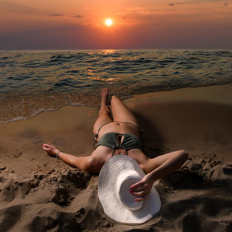 A woman in a sun hat lying in the sand at the water’s edge at sunset