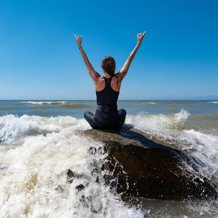 A woman in a yoga pose on a rock as a wave breaks at Oval Beach