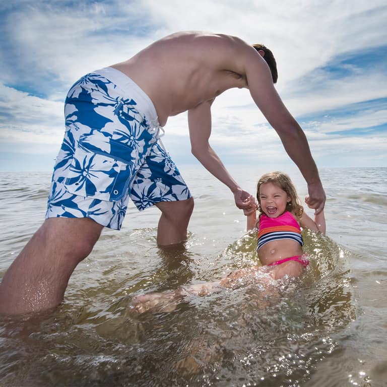 A father lifting his young daughter through a wave at Oval Beach
